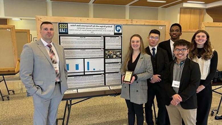 Penn State Altoona students Jazzmine McCauley, Ziwei (Will) Lin, Ray Ncube, Lam Vo, and Katelyn Kurtz standing next to their first-place poster, “Building Bridges with Pennsylvania State Police: An Assessment of Community Outreach,” and Trooper Joe Dunsmore, keynote speaker of the 2024 PACJE conference.  